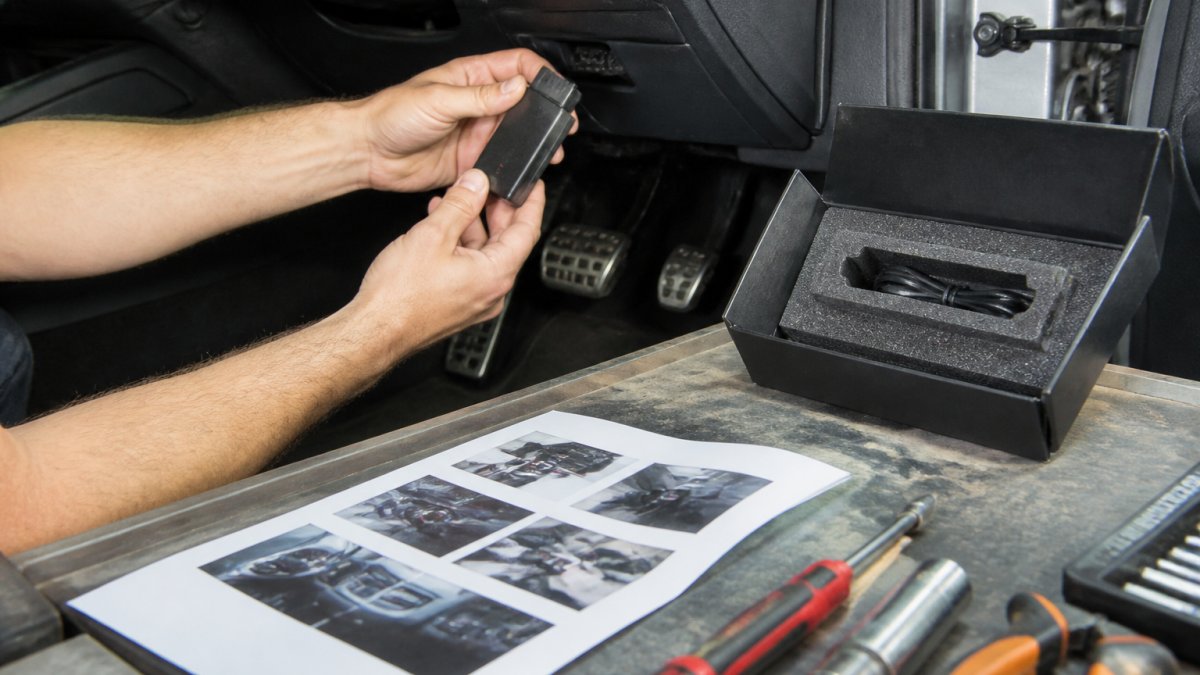 Technician's hands positioning a mileage blocker module near an OBD-II port under a vehicle dashboard, with a printed fitting guide and tools arranged on a workbench nearby