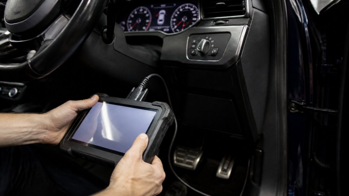 A mechanic's hands holding a diagnostic tablet connected to a car's OBD port, with the digital instrument cluster glowing in the background showing mileage readings