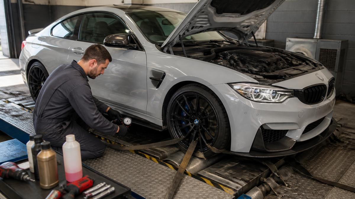 A mechanic in overalls checking tyre pressures and fluid levels on a performance car strapped to a rolling road in a workshop, with diagnostic laptop nearby